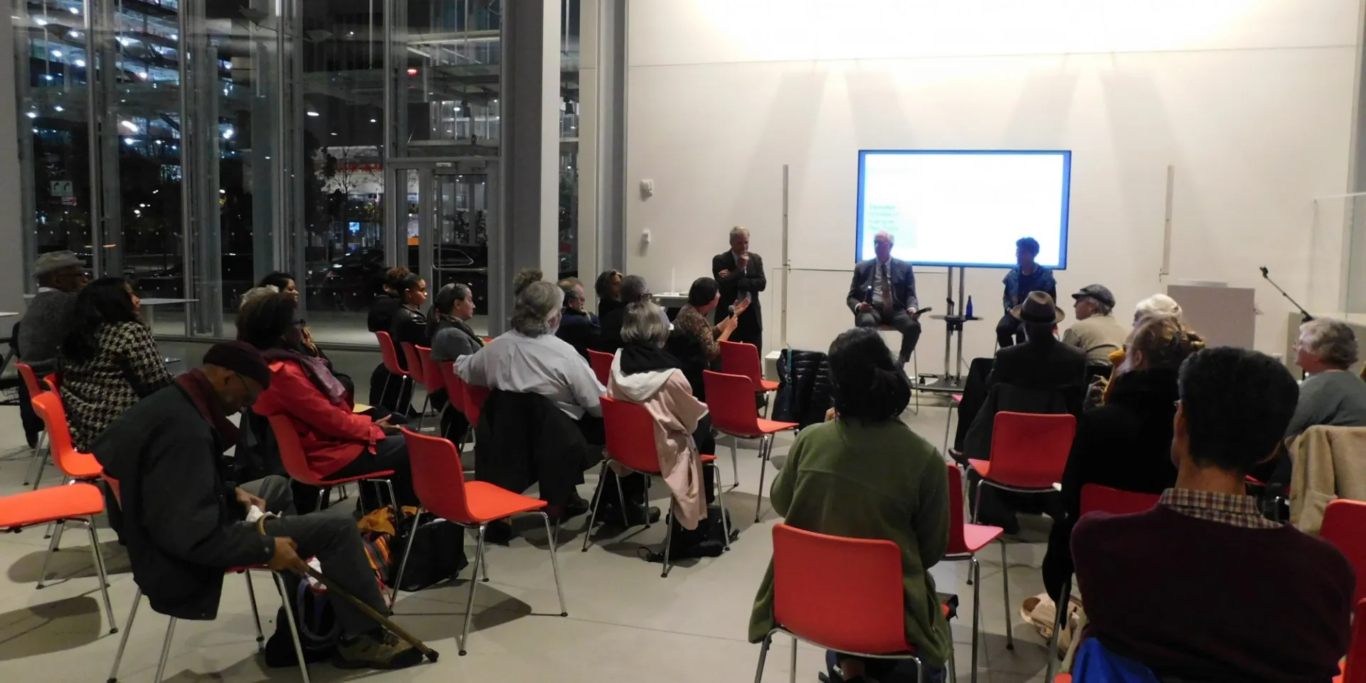 2 dozen people sitting on red chairs listening to 3 pannelists